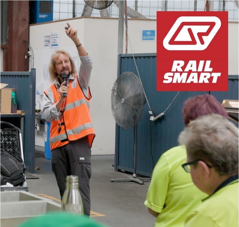 A Community Education Coordinator in a safety vest presenting to a group in a warehouse setting